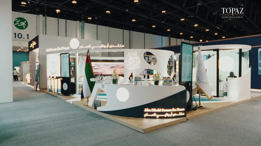 Curved white Abu Dhabi Forum for Peace exhibition stand with UAE flag and illuminated fascia at the Abu Dhabi International Book Fair