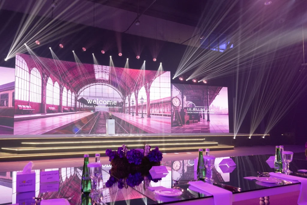 Stage with a railway-station LED backdrop, beam lighting and a purple floral centrepiece at Shjseen 2026
