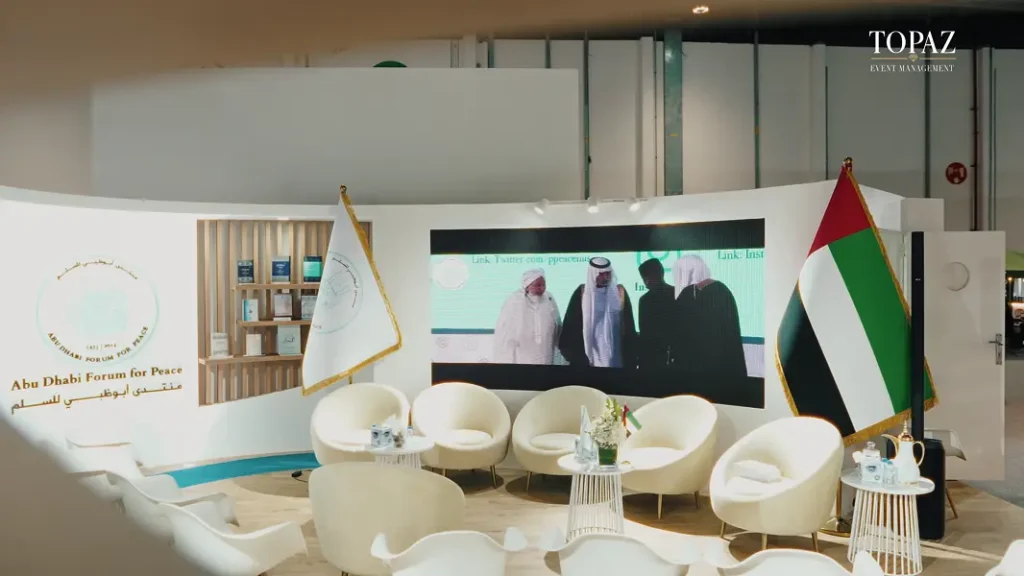 Lounge with LED screen and flags on the Abu Dhabi Forum for Peace exhibition stand at the Abu Dhabi International Book Fair