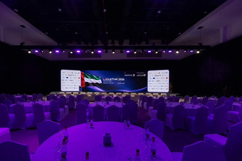 Wide view of the summit hall set with banquet tables under purple lighting, facing the LED stage