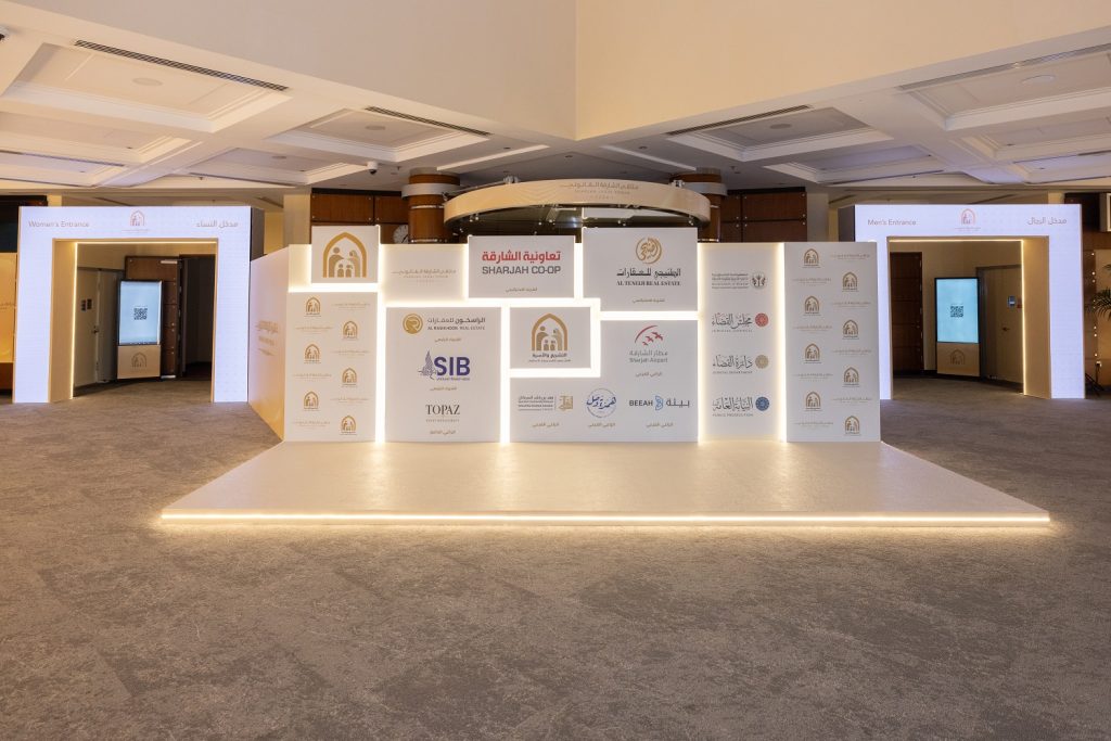 Illuminated white exhibition structure with sponsor logos and digital screens at the forum entrance