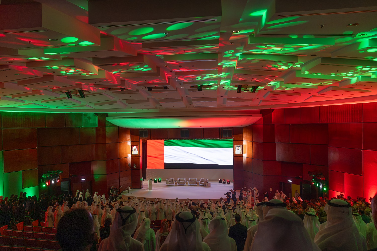 Audience in the main hall under green and red stage lighting with the UAE flag on screen