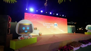 An outdoor evening stage setup for the Aisha Oasis for Innovation 2026 festival by the UAE Ministry of Economy. A large LED backdrop displays a vibrant orange and pink gradient with the event title in Arabic and English. In the foreground, a large, friendly robot mascot cutout with a digital smiling face stands on a lime-green platform. The stage is framed by spotlights, decorative flower beds, and palm leaves, organized by Topaz Events.