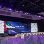 Angled view of the main summit stage and hall setup by Topaz Events at Grand Hyatt Dubai for the Quantum Innovation Summit 2026, showing round banquet tables, white VIP armchairs, overhead stage lighting, and the large LED backdrop displaying UAE flag graphics and sponsor walls.