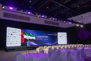 Angled view of the main summit stage and hall setup by Topaz Events at Grand Hyatt Dubai for the Quantum Innovation Summit 2026, showing round banquet tables, white VIP armchairs, overhead stage lighting, and the large LED backdrop displaying UAE flag graphics and sponsor walls.
