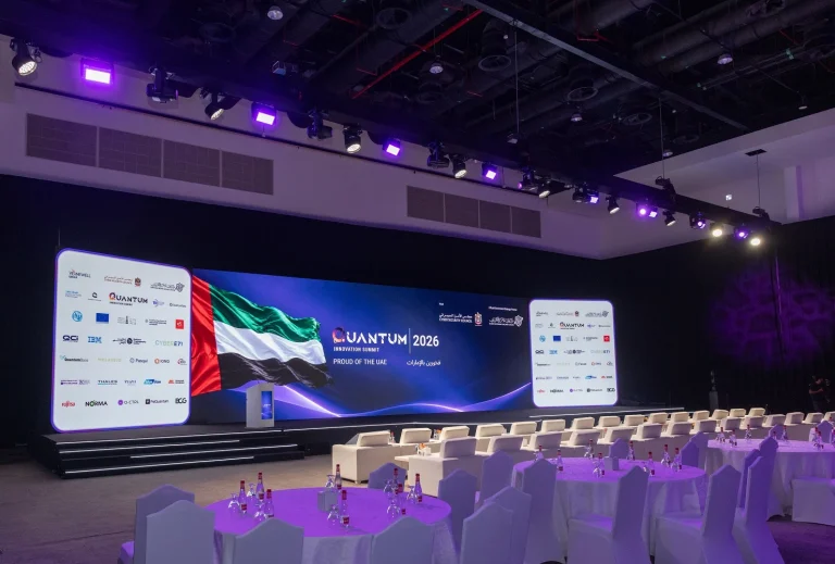 Angled view of the main summit stage and hall setup by Topaz Events at Grand Hyatt Dubai for the Quantum Innovation Summit 2026, showing round banquet tables, white VIP armchairs, overhead stage lighting, and the large LED backdrop displaying UAE flag graphics and sponsor walls.