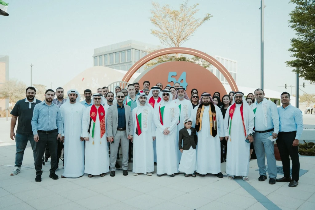 Sharjah Asset Management team group photo with UAE flag scarves under the 54 arch at the National Day event